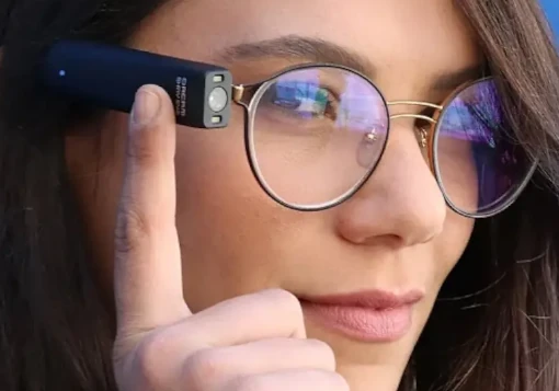 Senhora utiliza o Leitor Orcam MyEye encaixado na haste dos óculos. Aponta para indicar a leitura